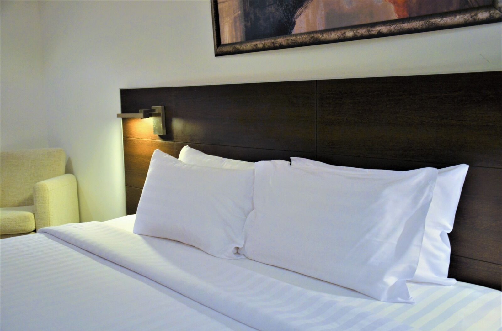 A neatly made bed with white linens and pillows, a dark headboard, and a wall-mounted light fixture.