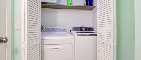 Een wasserette met een witte wasmachine en droger, een plank met een mand, een groene fles en een blauwe bak.