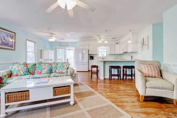 A spacious living room with a sofa, a coffee table, and a chair. There is a kitchen area with cabinets and a refrigerator in the background.