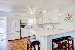A modern kitchen with white cabinets, a large island with stools, a ceiling fan, and a window providing natural light.