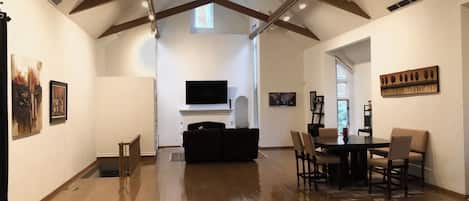 A spacious living area with a high ceiling, wooden beams, a fireplace, a flat-screen TV, and a dining area with chairs and a table.
