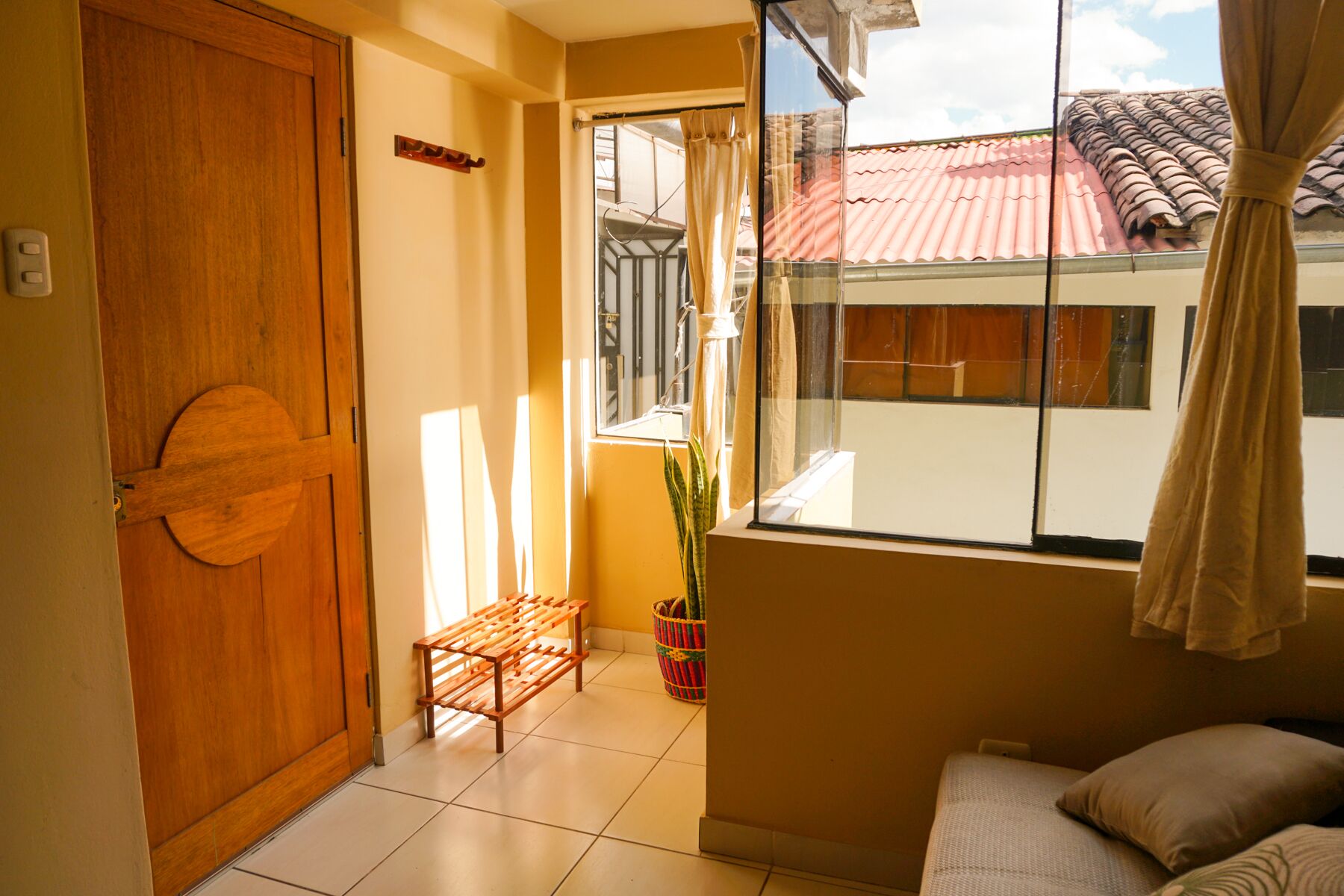A room with a wooden door, a bench, a potted plant, a bed, and a window with a view of a tiled roof.