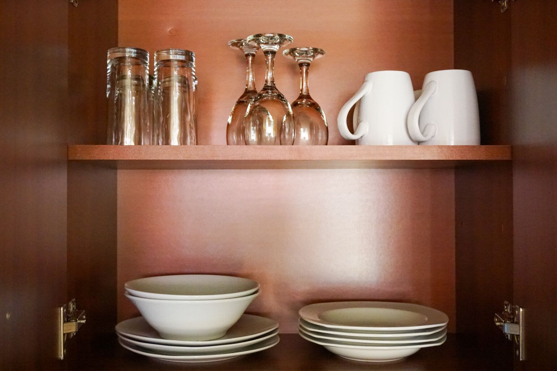 A wooden cabinet with shelves holding glass bottles, wine glasses, and white mugs. Below, there are stacked white bowls and plates.
