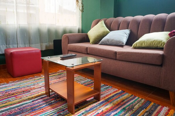 A living room with a brown sofa, a glass coffee table, a red ottoman, and a colorful striped rug.