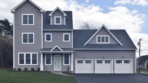 A two-story house with a front porch, a garage with two doors, and a small front yard. - Summer Oasis in Narragansett! 5BR Home Perfect for Family & Friends! (Narragansett)