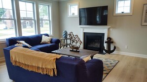 A living room with a blue sofa, a fireplace, a TV, and a view of the outside. - Summer Oasis in Narragansett! 5BR Home Perfect for Family & Friends! (Narragansett)