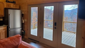 A cabin interior with a wooden ceiling, a refrigerator, a couch, and a large sliding glass door leading to a deck with a view of autumn trees. - Romantic Getaway! Hot Tub & Mountain Views, King Bed (whittier)