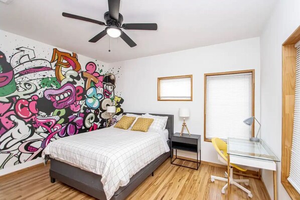 A bedroom with a bed, a desk, a chair, a ceiling fan, and a wall with colorful graffiti.