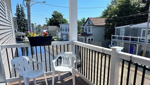 Una terraza de madera con barandas blancas, dos sillas de plástico blanco y una jardinera con flores.
