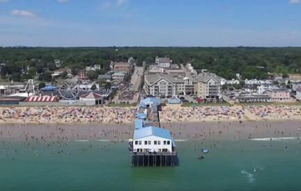 Um píer se estende sobre a água, com uma praia e edifícios ao fundo.