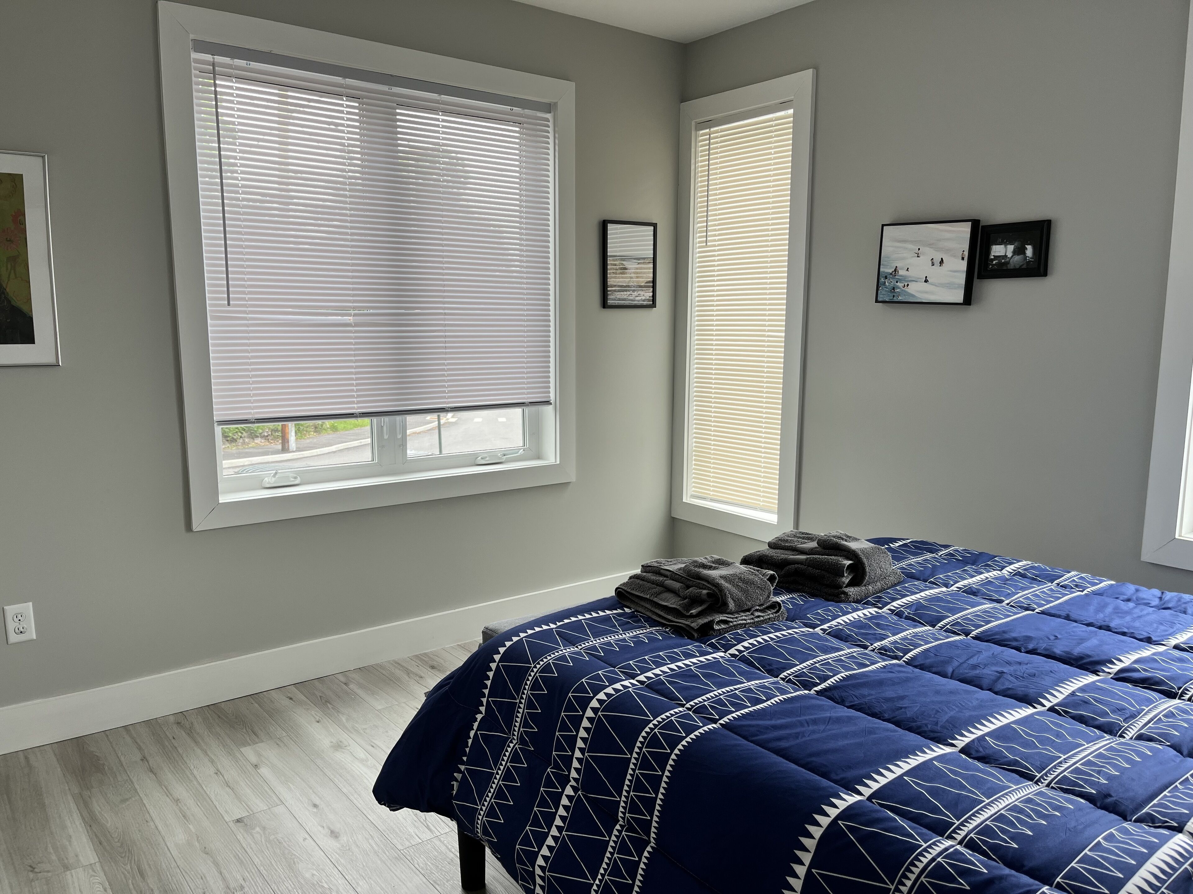 A bedroom with a bed, two windows with blinds, and a wall with framed pictures.