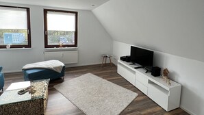 A modern living room with a sloped ceiling, a television on a white console, a blue sofa, and a glass coffee table.