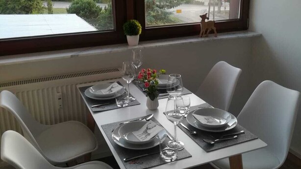 A dining table set for four with white chairs, a potted plant, and a small wooden deer figure on the windowsill.