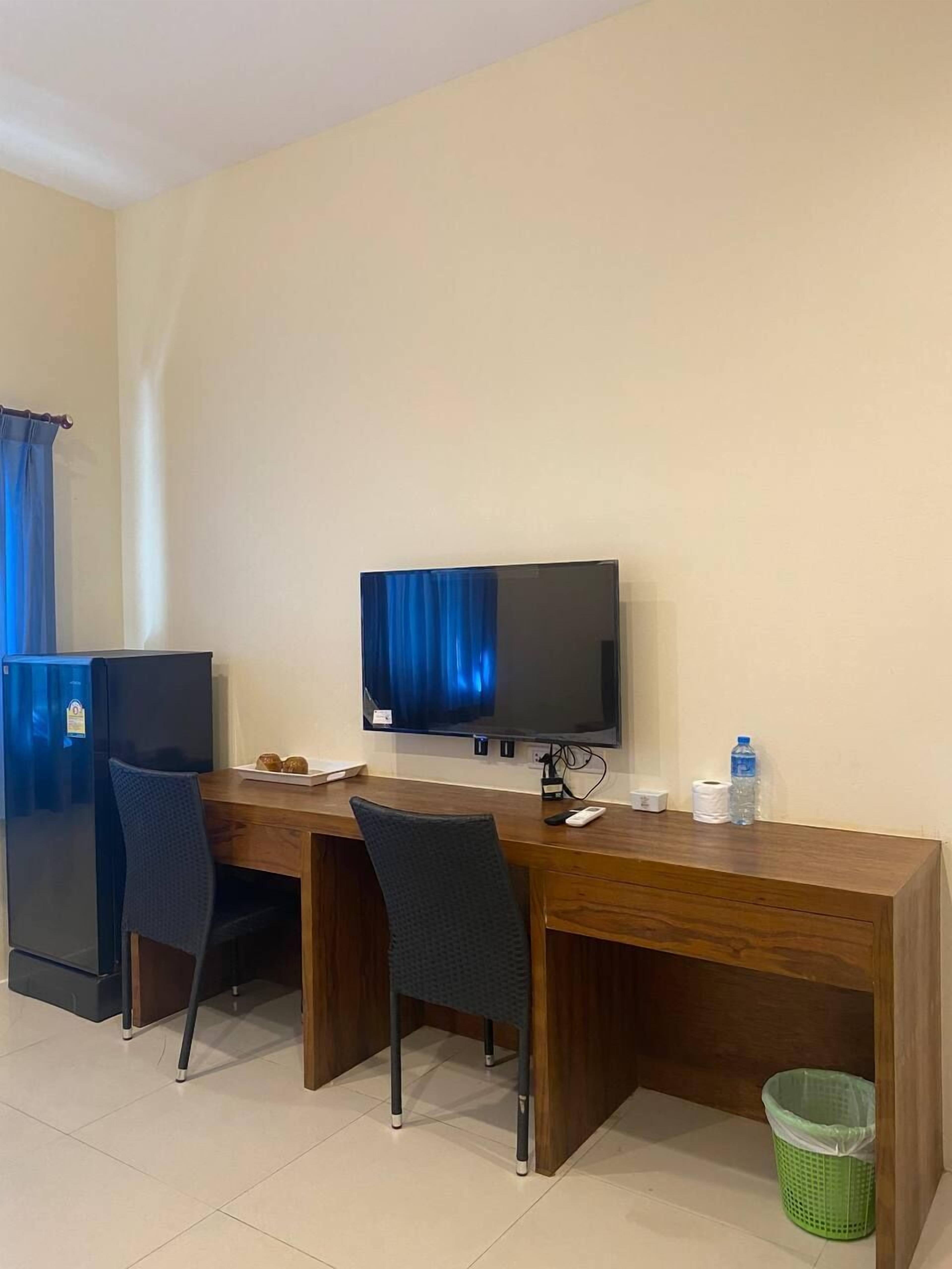 A hotel room with a wooden desk, two chairs, a television, a refrigerator, a trash bin, and a water bottle.