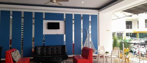 Un hall con un sofá de cuero negro, dos sillones rojos, un ventilador de techo y un televisor montado en la pared.