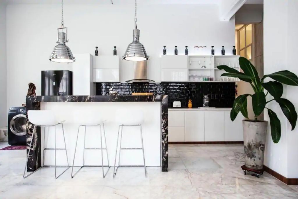 Een moderne keuken met een marmeren aanrecht, witte barkrukken, hanglampen en een potplant.
