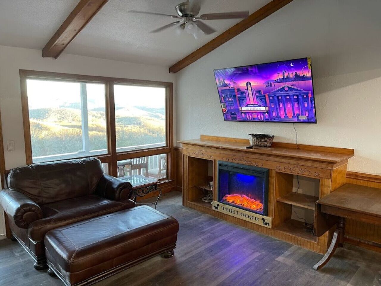 A living room with a leather sofa, a fireplace, a television mounted on the wall, and a view of the outdoors through large windows.