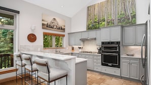 A modern kitchen with a central island, bar stools, and a large window with a view of trees. - Beautiful Mountain Home With Hot Tub on Free Bus Route (Vail)