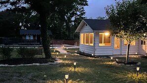 Une petite maison de plain-pied, dotée d’un toit en pente et de grandes fenêtres, entourée d’une cour verdoyante et agrémentée de plusieurs lampadaires.