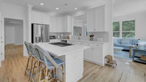 A modern kitchen with white cabinetry, a central island with bar stools, and a dining area with a light-colored wood floor. - Award-Winning Resort Amenities | Fairway Oaks 33 (Isle of Palms)