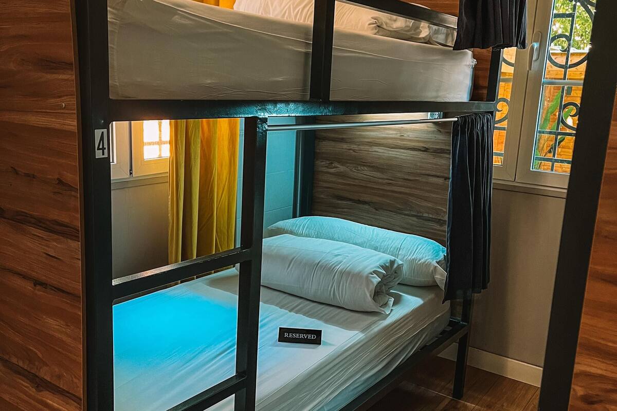 Quarto com beliches, um aviso de 'RESERVED' na cama inferior, paredes de madeira e uma janela com cortinas amarelas.