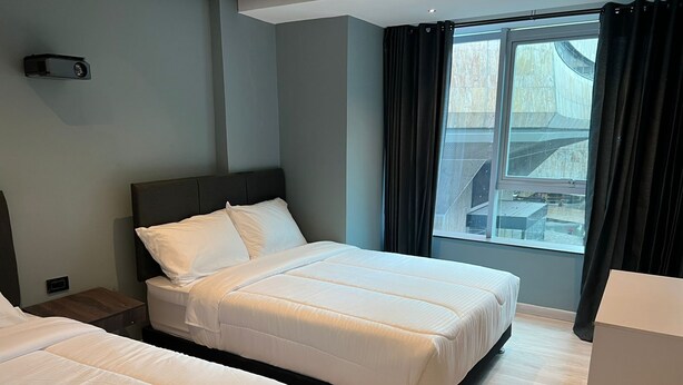 Habitación de hotel con dos camas, un ventanal grande con cortinas, una mesita de noche y un armario pequeño.