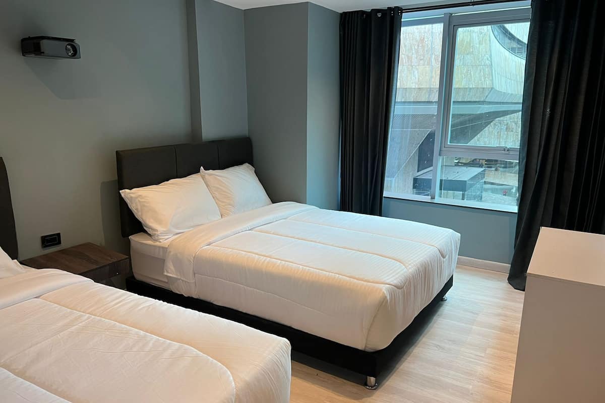 Habitación de hotel con dos camas, un ventanal grande con cortinas, una mesita de noche y un armario pequeño.
