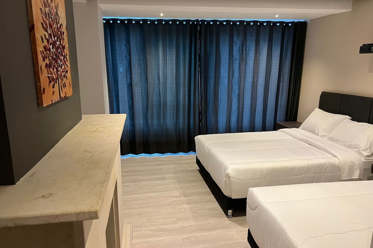 Habitación de hotel con dos camas, un escritorio y un amplio ventanal con cortinas azules.