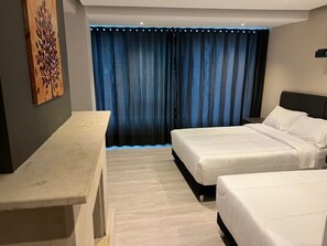 Habitación de hotel con dos camas, un escritorio y un amplio ventanal con cortinas azules.