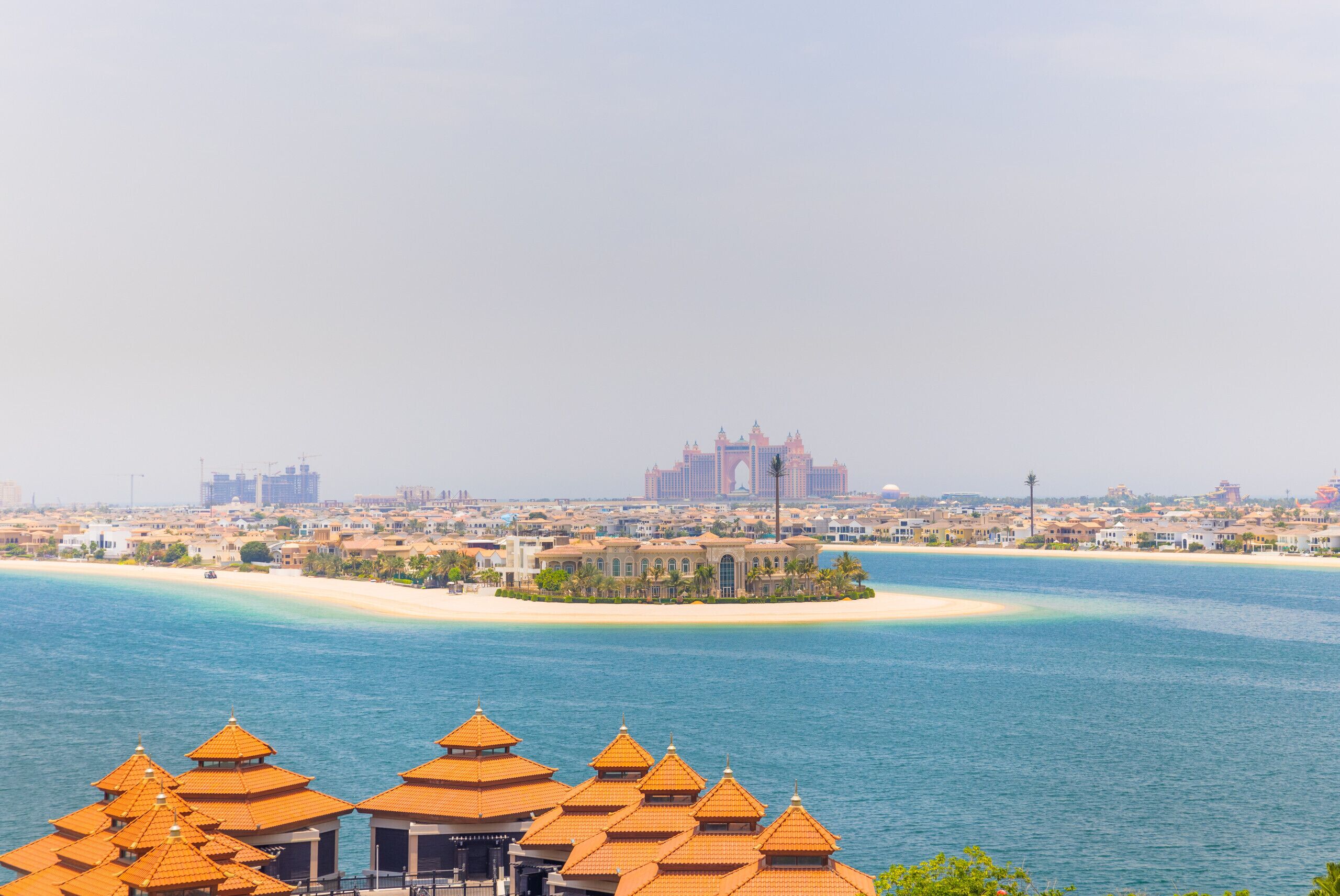 A coastal resort with multiple buildings featuring traditional architecture, a sandy beach, and a view of a distant cityscape.