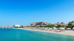 Un hotel frente a la playa con varias edificaciones, una playa de arena y sombrillas.