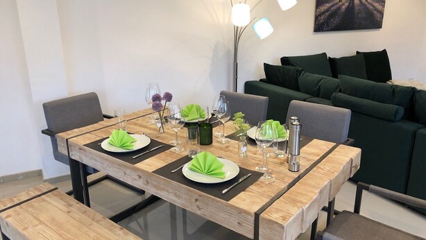 Uma área de refeições com mesa de madeira posta para dois, um banco e um sofá ao fundo.