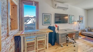 A room with a wooden desk, two chairs, a window with a view, an air conditioning unit, and artwork on the walls. - The Brick Unit B on F st Downtown Salida! (Salida)