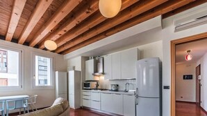 A compact kitchen with white cabinets, a refrigerator, and a microwave. There is a dining area with a table and chairs, and a sofa. The room has wooden beams on the ceiling and a window with blinds.