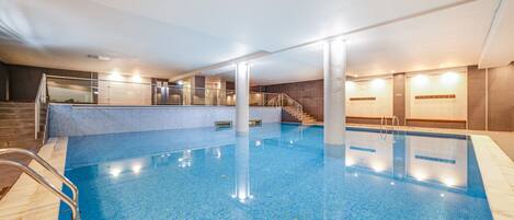 An indoor swimming pool with clear water, a metal ladder, and a staircase leading to a higher level.