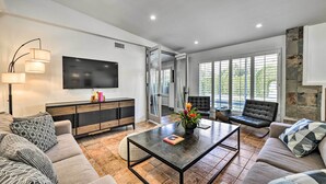 Un salon moderne avec un téléviseur à écran plat, une table basse et un canapé. Il y a une grande fenêtre avec des stores, un mur d’accent en pierre et une porte qui donne sur une autre pièce.