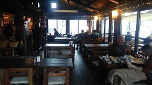 A rustic restaurant interior with wooden tables and benches, string lights, and large windows offering a view of the outdoors. - Kulindağ Dağ Evi (Beykoz)