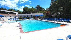 A swimming pool with blue lounge chairs and umbrellas, surrounded by trees and a building. - Seawinds II (Dennis Port)