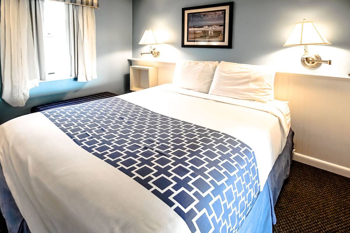 A hotel room with a bed, two bedside lamps, a framed picture on the wall, and a window with curtains.