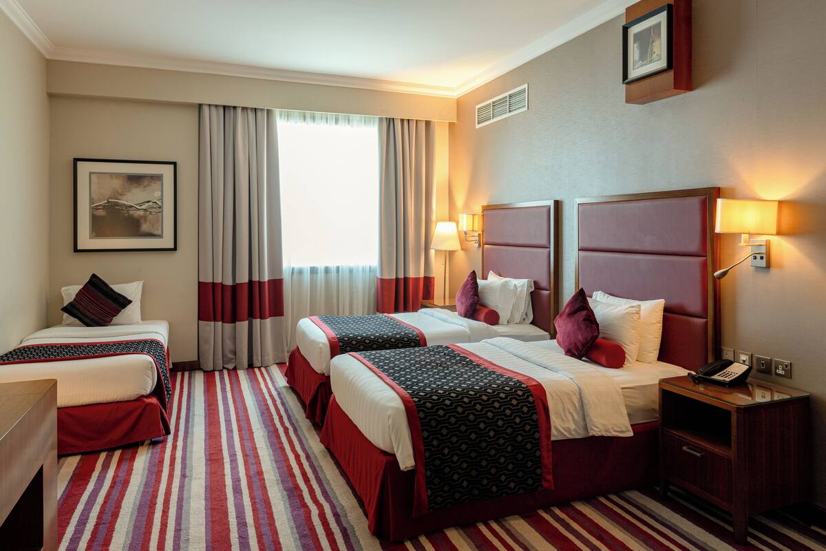 Une chambre d’hôtel avec deux lits, une grande fenêtre avec des rideaux, une table de chevet avec une lampe et un tableau encadré au mur.