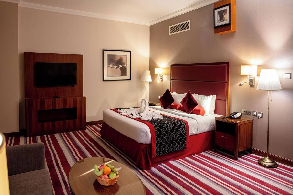 Une chambre d’hôtel équipée d’un lit, d’une table de chevet, d’une lampe, d’une télévision et d’un panier de fruits.