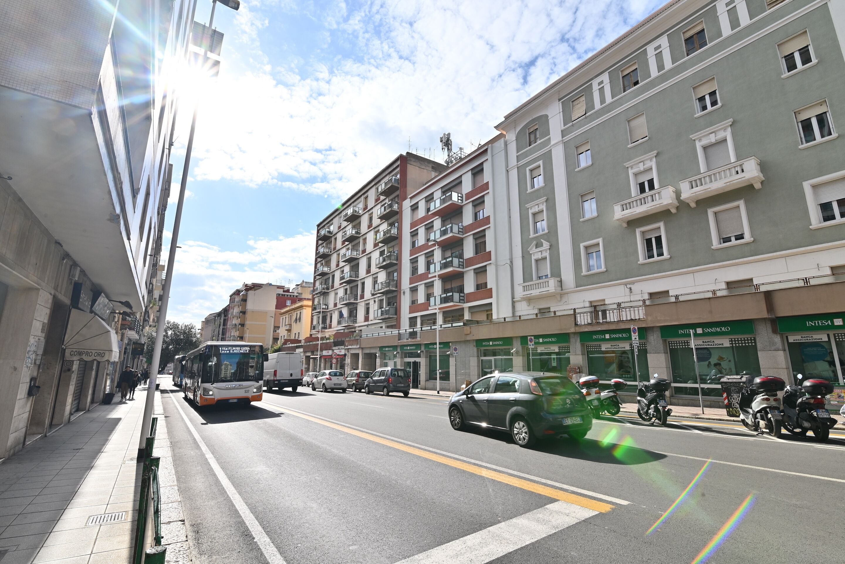 A multi-story building with balconies, a street with cars and a bus, and storefronts with signage.