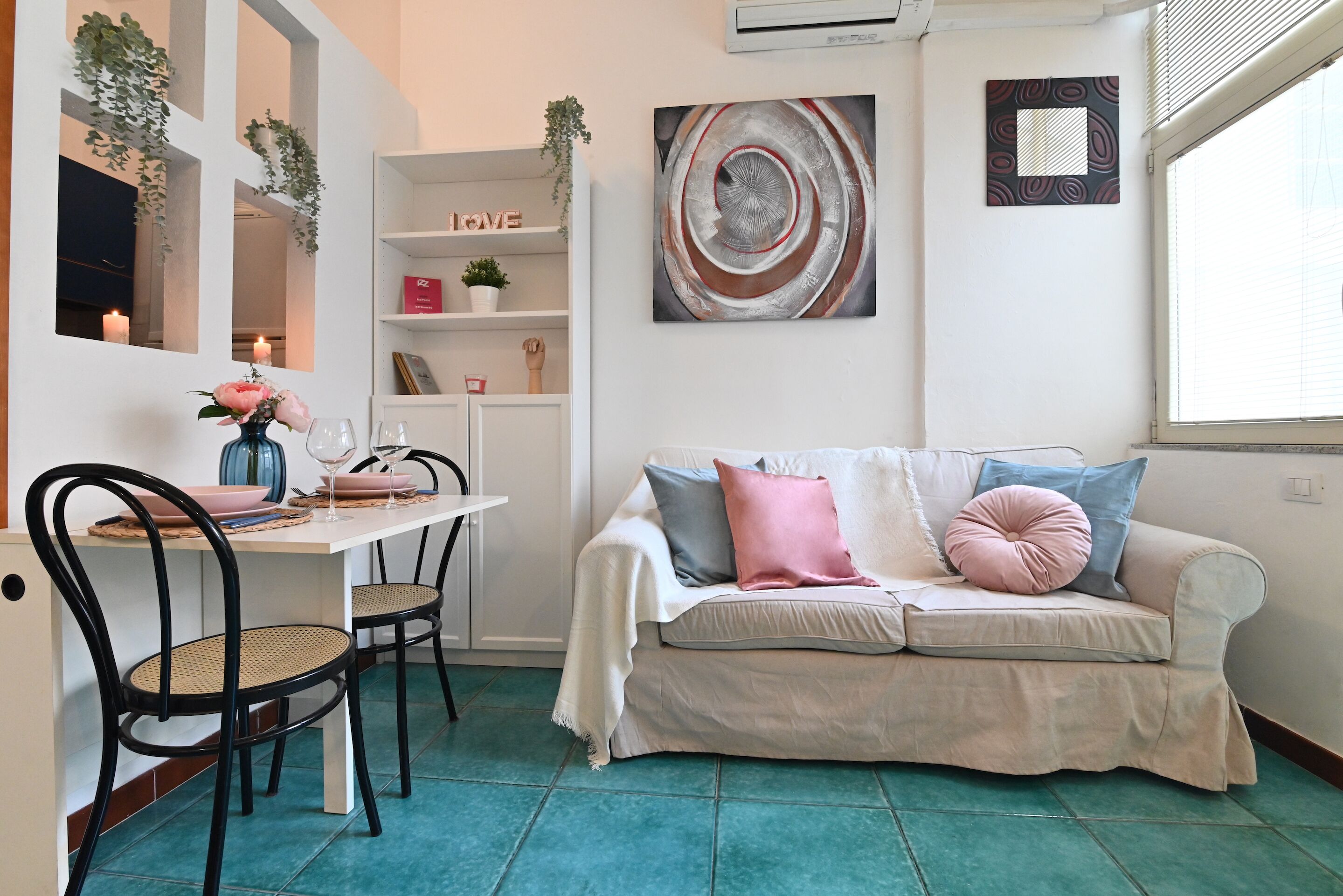 A cozy living area with a white sofa, teal floor tiles, a dining table with chairs, and a bookshelf with decorative items.