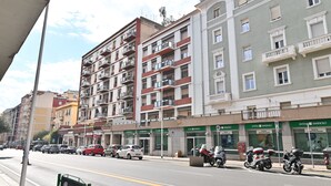 A multi-story building with balconies, a street with parked cars and motorcycles, and storefronts with signage. - IzzHome City Love (Cagliari)