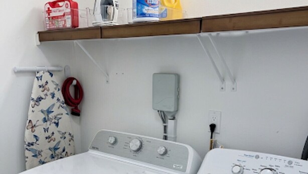 A laundry area with two white washing machines, a red iron, a bottle of detergent, and a hanging iron on a white wall.