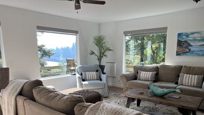 Un salón con sofá, sillón y mesa de centro. Ventanas ofrecen vistas a zonas verdes y una montaña.