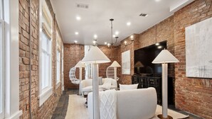 A modern room with exposed brick walls, a large bed, a seating area with chairs, and a flat-screen TV.