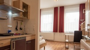 Una cucina moderna con mobili in legno, forno integrato e lavello. Si vede un letto con copriletto bianco e una finestra con tende rosse.
