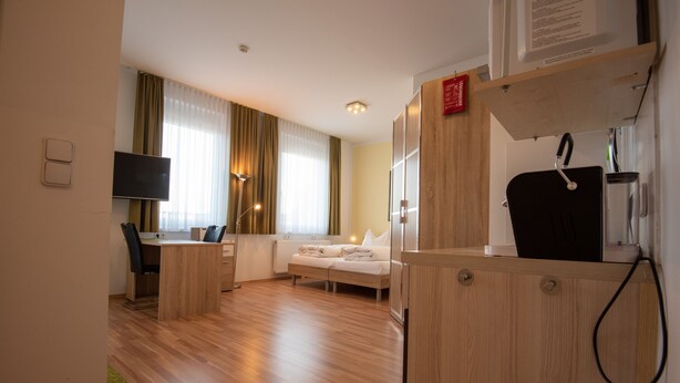 Una camera d'albergo con pavimento in legno, un letto, una scrivania, una sedia, un televisore e una finestra con tende.