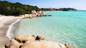 A coastal beach with clear turquoise water, large rocks, and a forested hillside.